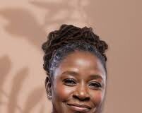 The Land Remembers: Tourism, Legacy, and the Haitian Future **Alt Text:** Portrait of a woman with medium-dark skin and neatly styled dreadlocks pulled up into a bun. She is wearing a bright red blazer and smiling warmly. The background is a soft beige with abstract shadows of leaves, giving a calm and elegant ambiance.