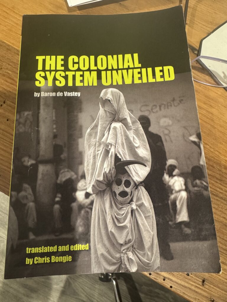 **Alt text:** Cover of the book *The Colonial System Unveiled* by Baron de Vastey, translated and edited by Chris Bongie. The cover features a striking black-and-white photo of a person draped in a white shroud-like garment, holding a painted mask resembling a skull. The background shows a group of similarly dressed figures and a wall with the word "Senate" faintly visible. The title appears in bold yellow-green text at the top.