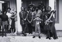 Friendship in the Age of Self-Improvement Black and white photograph of a large, diverse group of people gathered in front of a decorated home, standing on a porch with two large columns. The group includes men, women, and children of various ages, dressed in a mix of formal and casual attire. Some individuals are embracing or holding hands, while others stand side by side, smiling at the camera. A young child in the front playfully makes binocular shapes with their hands. The background features a festive entrance with wreaths on the doors, suggesting a holiday or celebratory gathering. The atmosphere conveys warmth, connection, and togetherness.