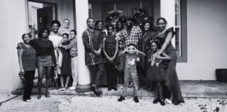 Friendship in the Age of Self-Improvement Black and white photograph of a large, diverse group of people gathered in front of a decorated home, standing on a porch with two large columns. The group includes men, women, and children of various ages, dressed in a mix of formal and casual attire. Some individuals are embracing or holding hands, while others stand side by side, smiling at the camera. A young child in the front playfully makes binocular shapes with their hands. The background features a festive entrance with wreaths on the doors, suggesting a holiday or celebratory gathering. The atmosphere conveys warmth, connection, and togetherness.