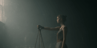 Part II — The Weight Bent by Nostalgia A dark, moody, and dramatic digital painting of a woman in a long, tattered dress standing in a dimly lit, dusty interior. She holds up a large, antique balance scale or weighing pan filled with rough, heavy rocks or rubble, illuminated by a single shaft of light from an unseen window.